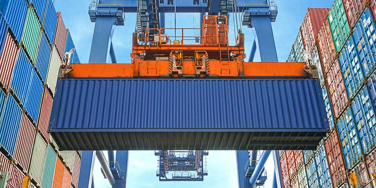 Services - Shipping and Logistics - a photo of a crain lifting an ocean container onto a large wall of colorfull containers in a shipping yard
